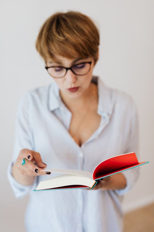 Free From above pensive female with short hair wearing casual light blue shirt and eyeglasses reading paper book in hardcover while standing against white plain wall Stock Photo