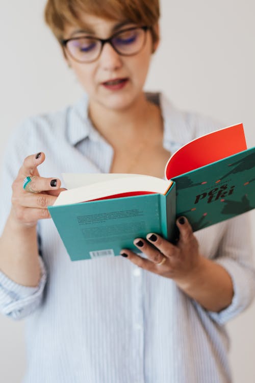Free Crop positive female in casual shirt and eyeglasses reading interesting novel in hardcover while standing against white background Stock Photo
