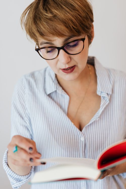 Free Crop calm female in eyeglasses and casual light blue shirt absorbedly reading thick book in hardcover against white wall Stock Photo