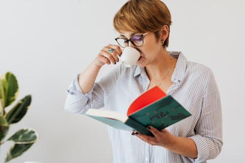 Crop pensive female in eyeglasses and casual wear reading interesting book in hardcover and drinking hit aromatic coffee from white ceramic cup against white wall and green plant