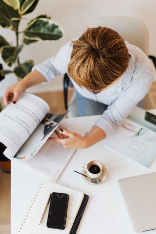 Free From above anonymous female in jeans and light blue shirt working at desk with gadgets and papers while turning over magazine and having cup of hot americano Stock Photo