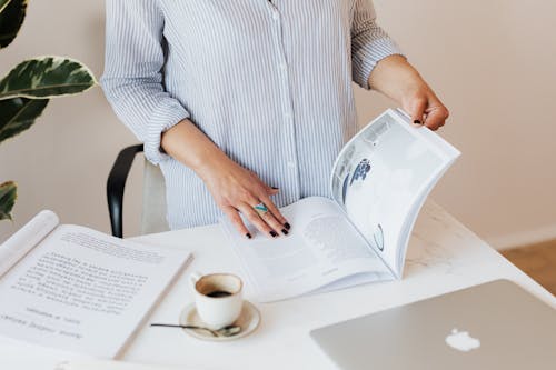 Free Crop unrecognizable female in light blue shirt standing near desk with laptop and opened book while reading professional journal and enjoying cup of aromatic coffee Stock Photo