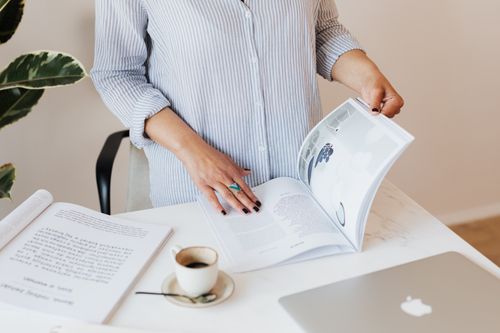 Free Crop unrecognizable female in light blue shirt standing near desk with laptop and opened book while reading professional journal and enjoying cup of aromatic coffee Stock Photo