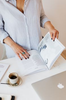 上图中，一位身穿蓝色衬衫的匿名女性站在办公桌旁，手里拿着笔记本电脑和一杯刚冲好的咖啡，正对着白墙翻阅专业日记