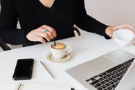 Crop unrecognizable female in black clothes working on modern netbook and stirring cup of fresh aromatic espresso