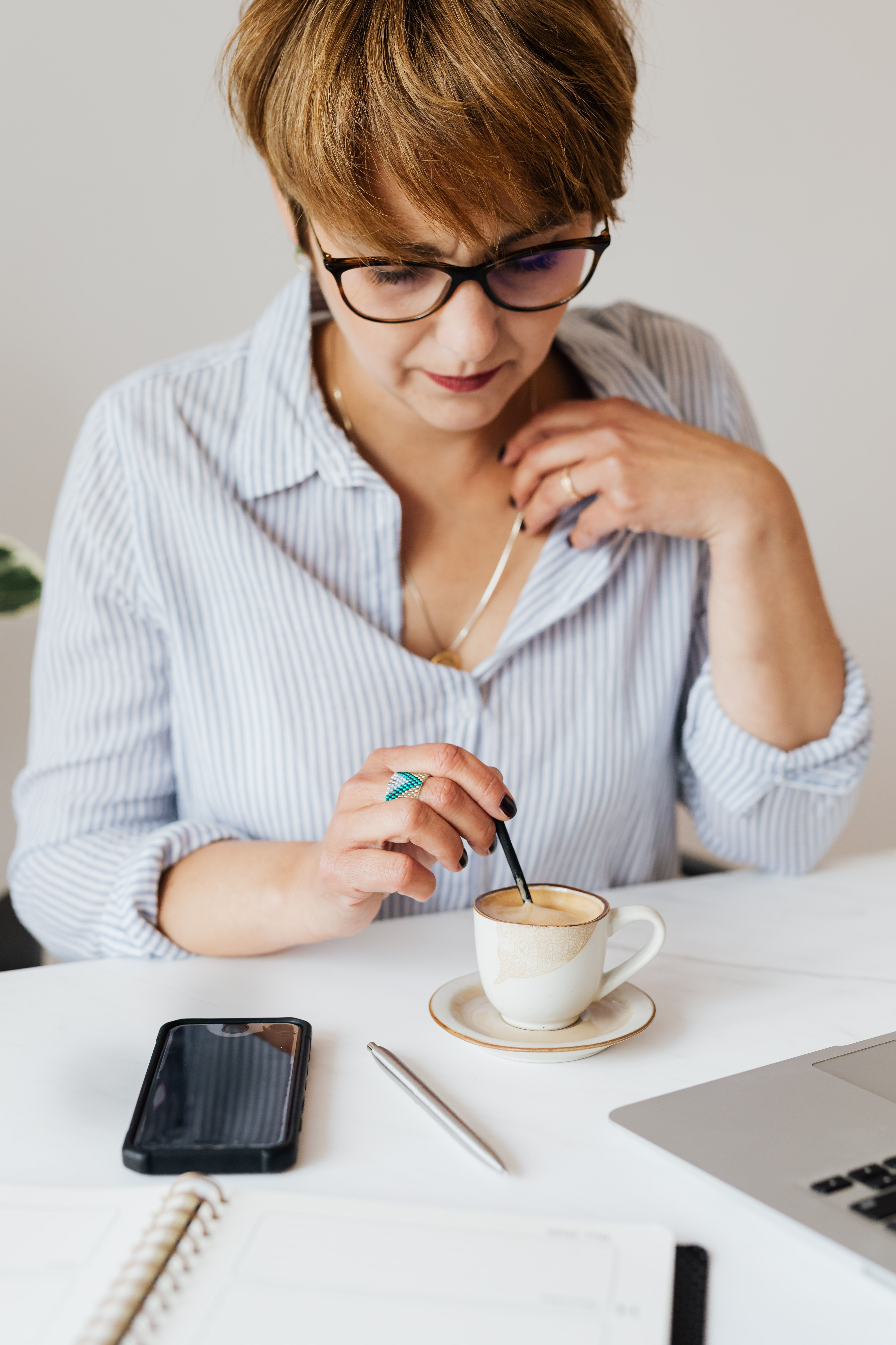 Crop serious female in eyeglasses and formal shirt sitting at table with gadgets and notebook while stirring aromatic fresh coffee