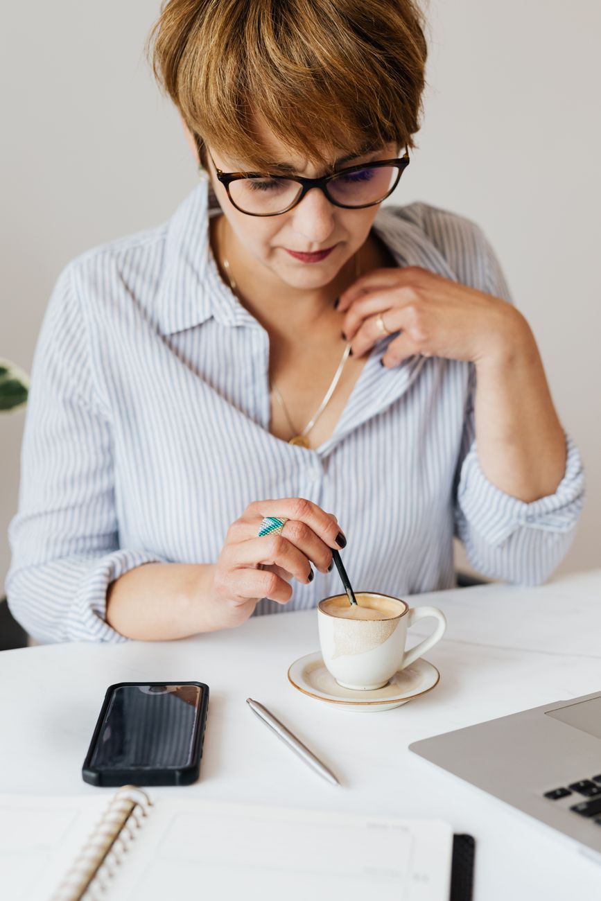 Crop serious female in eyeglasses and formal shirt sitting at table with gadgets and notebook while stirring aromatic fresh coffee