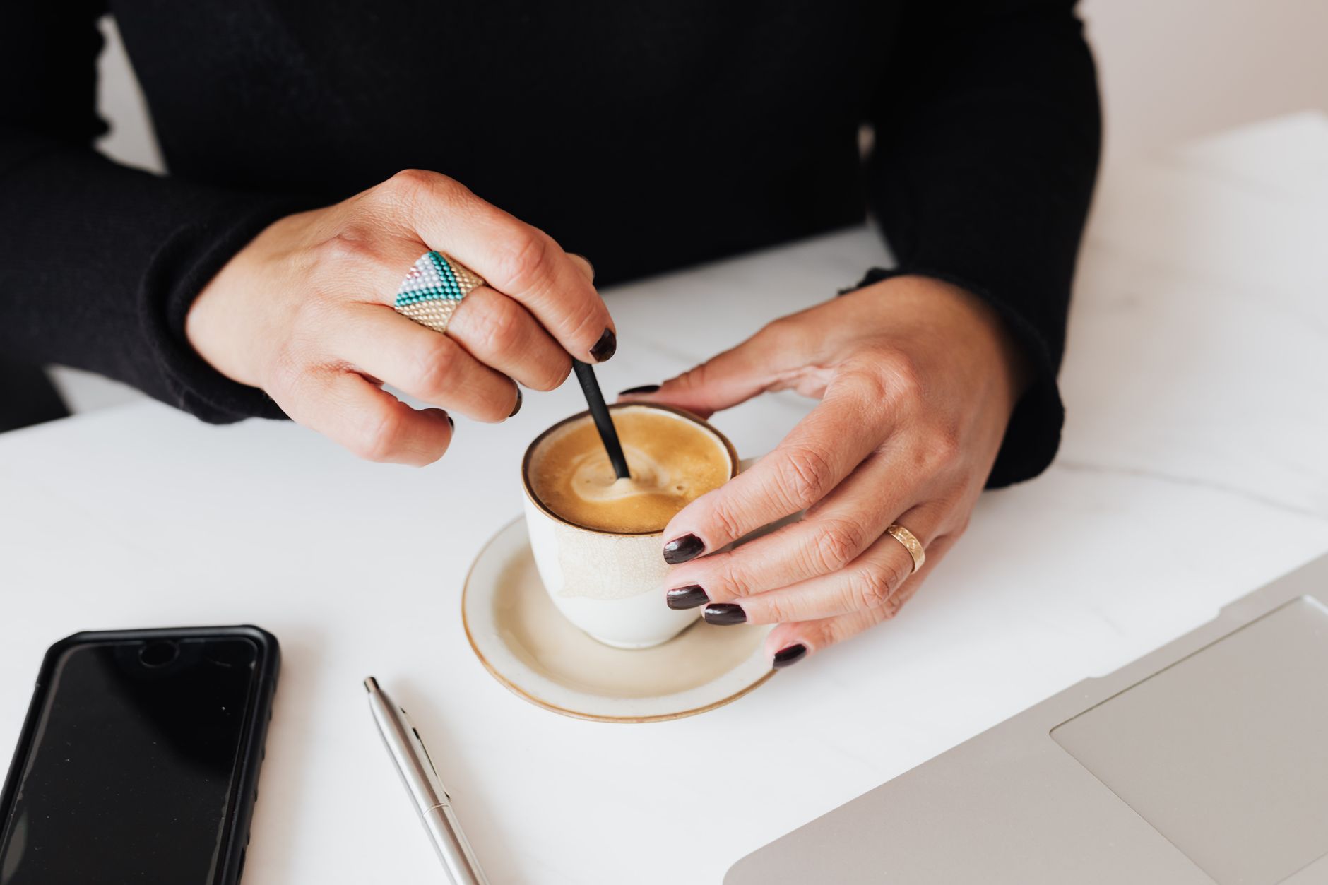 Close-up of a woman stirring espresso in a cup at a workspace with a smartphone and laptop nearby.