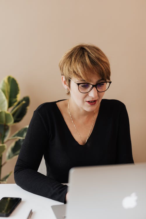 Content elegant adult female with short haircut in eyeglasses using laptop while working on business project in modern office