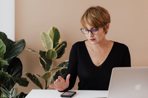 Free Concentrated female manager in casual outfit and eyeglasses reading message on smartphone while sitting at table with laptop in cozy workspace Stock Photo