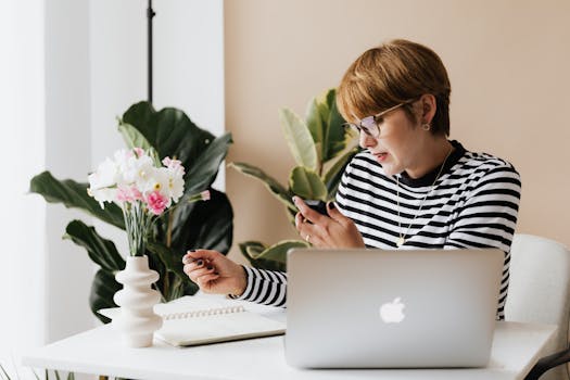 Focused woman using smartphone and laptop in modern home office with plants and notebook.