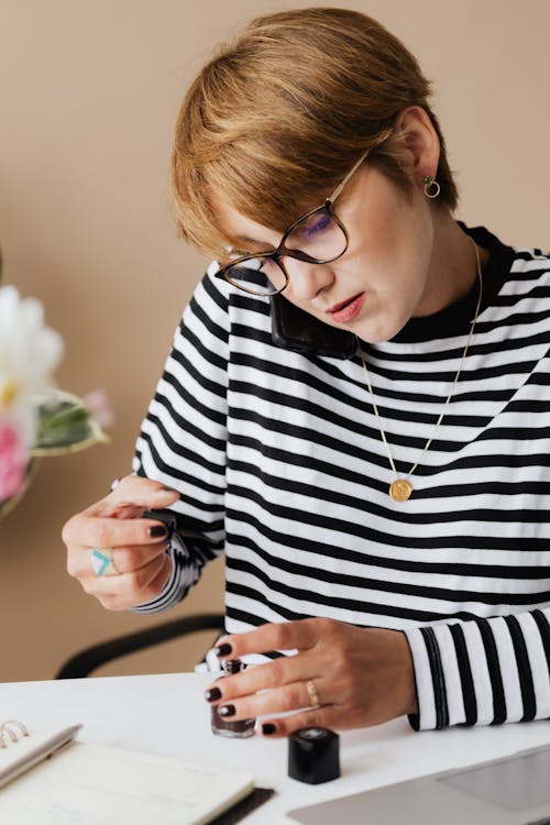 Free Attentive middle aged female in casual wear and eyeglasses applying nail polish and talking on smartphone while working remotely from home Stock Photo