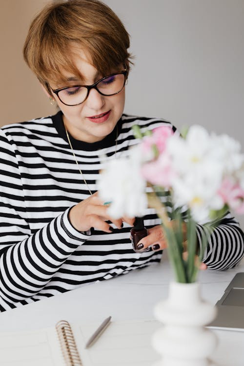 Free Young woman in glasses painting nails at home while sitting at table Stock Photo