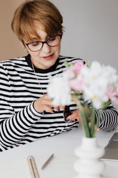 Stylish woman with glasses applying nail polish at her desk, surrounded by flowers and a laptop.
