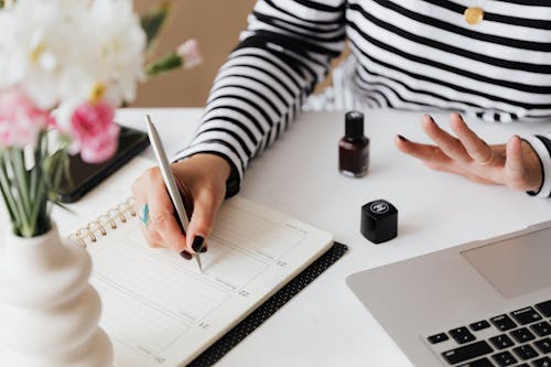 Free Unrecognizable female using pen and writing notes in notepad while sitting at table and waiting when polish nail getting dry Stock Photo