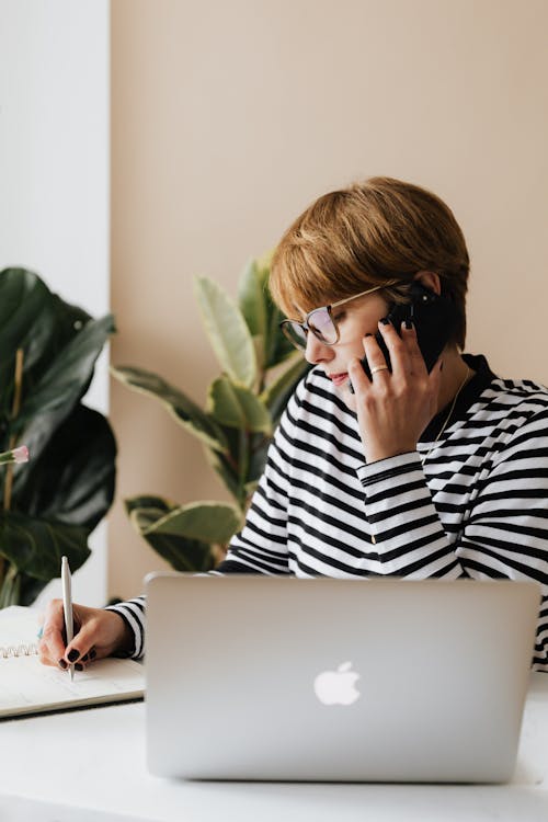 Concentrated female freelancer wearing casual clothes and eyeglasses sitting at table and having conversation on smartphone and writing notes in notepad while working with laptop