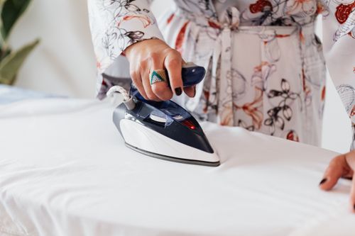 Free Crop unrecognizable female with manicured hands wearing classy silk robe and stylish ring ironing white sheets on ironing board Stock Photo
