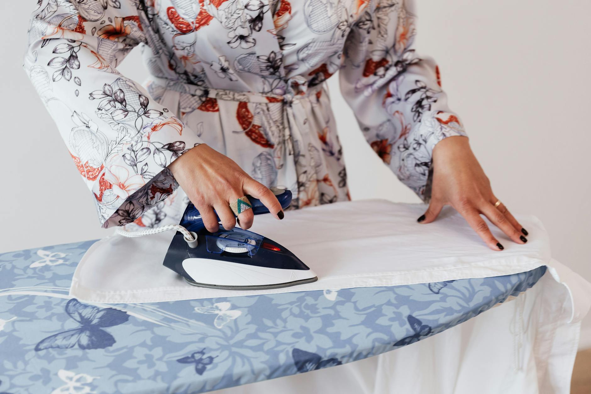 Crop faceless female in stylish silk robe ironing formal white shirt on blue ironing board by using contemporary small iron against white plain wall
