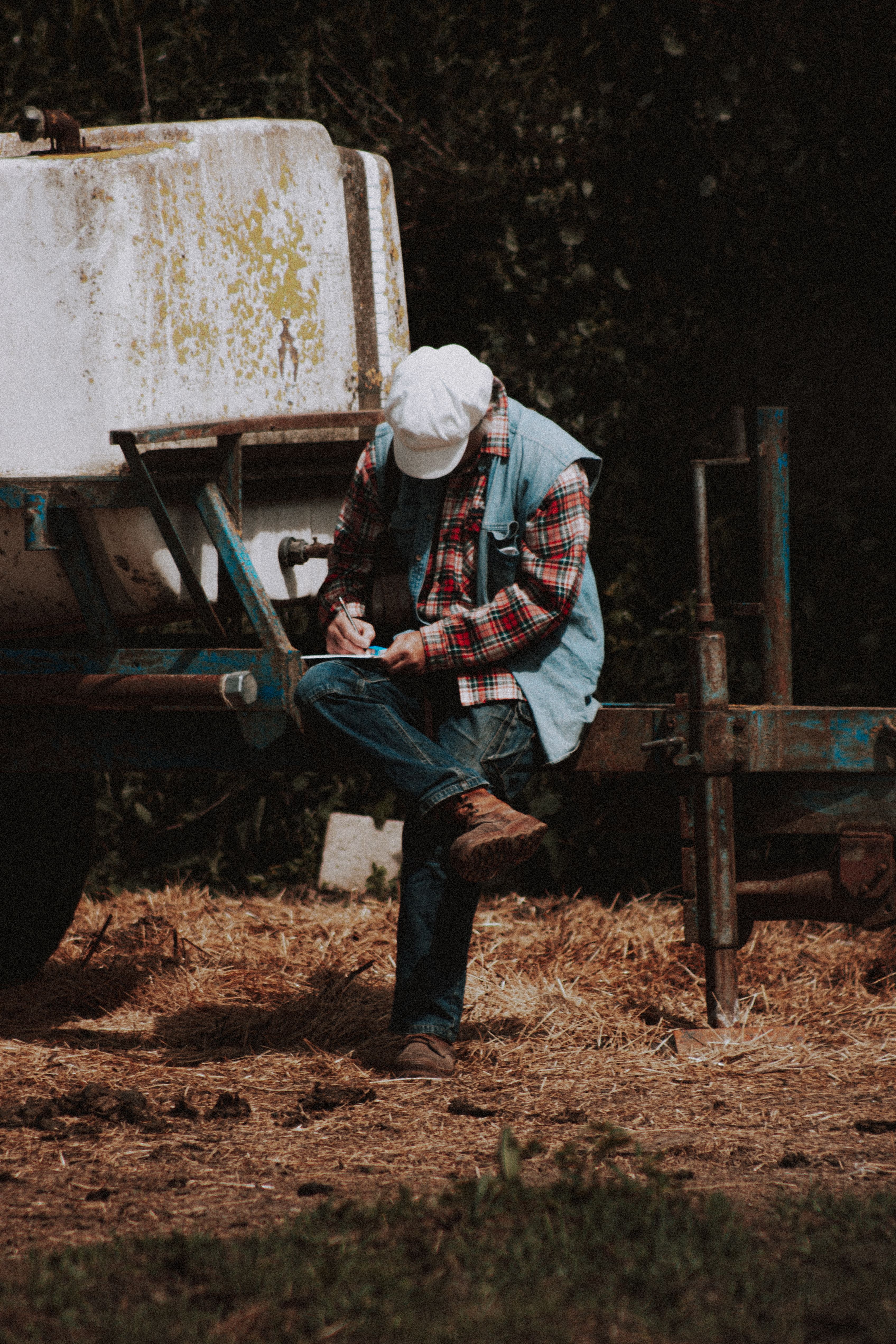Full body anonymous man in casual clothes and cap sitting on shabby trailer and writing in notepad during break in work on ranch
