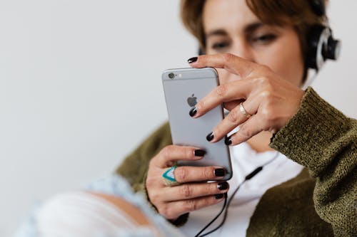 Free Crop blurred focused female in casual warm clothes with classy ring surfing contemporary cellphone while enjoying favorite music via wired headphones against white plain wall Stock Photo