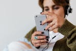 Crop blurred focused female in casual warm clothes with classy ring surfing contemporary cellphone while enjoying favorite music via wired headphones against white plain wall