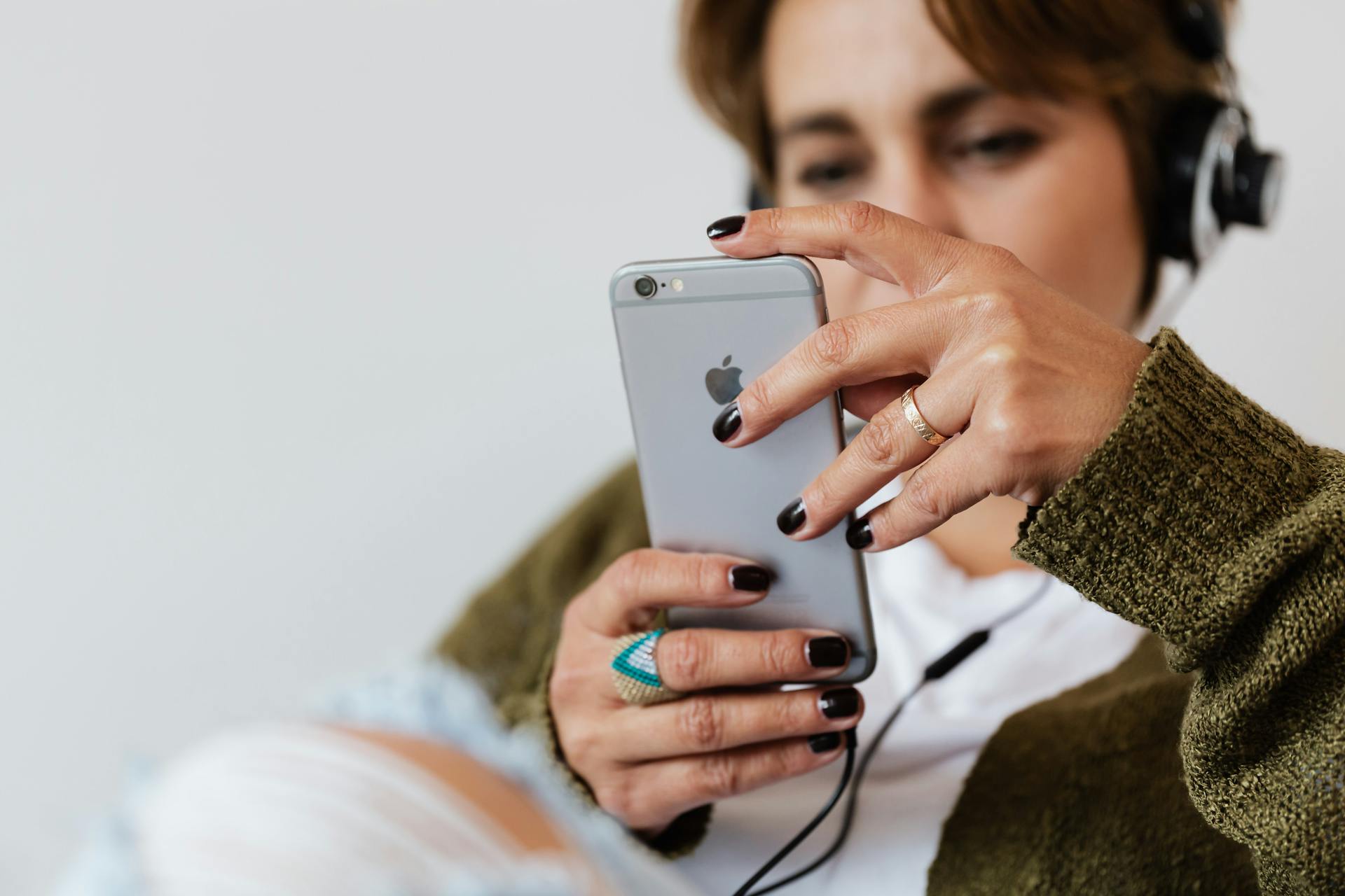 Crop blurred focused female in casual warm clothes with classy ring surfing contemporary cellphone while enjoying favorite music via wired headphones against white plain wall