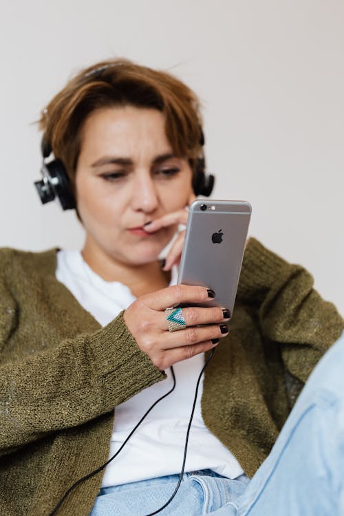 Free Pensive female in casual warm clothing sitting against white wall and touching face while browsing modern smartphone and listening to good music in wired headphones Stock Photo