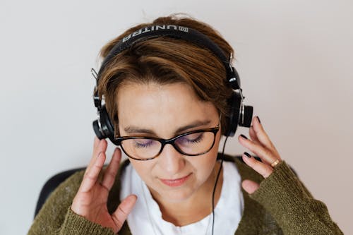 Free Positive relaxed female in casual warm jacket and eyeglasses resting against white wall with eyes closed and enjoying favorite music in wired headphones Stock Photo