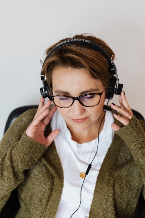 Glad relaxed female wearing eyeglasses and warm casual clothes resting on chair with eyes closed and listening to good music via wired headphones on white background