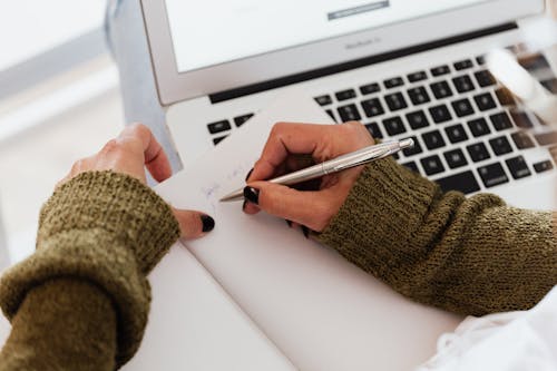 From above crop unrecognizable female in casual warm sweater taking notes in opened notebook with stylish silver pen while using modern netbook