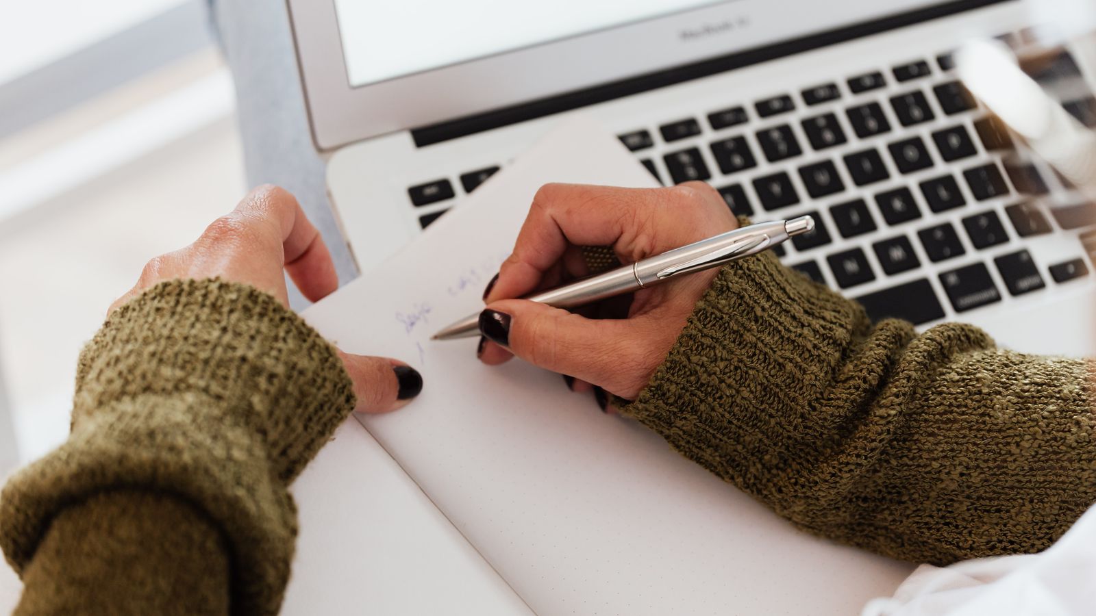 A woman taking notes beside an open laptop at a desk