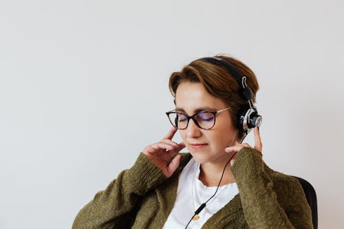 Free Content glad female wearing eyeglasses and headphones listening to good music and touching headset while sitting with eyes closed against white wall Stock Photo