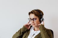 Content glad female wearing eyeglasses and headphones listening to good music and touching headset while sitting with eyes closed against white wall