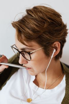一位专注的女性身着休闲服，戴着时尚的项链和眼镜，靠在白墙上，戴着耳机听播客，并用银笔轻触嘴唇