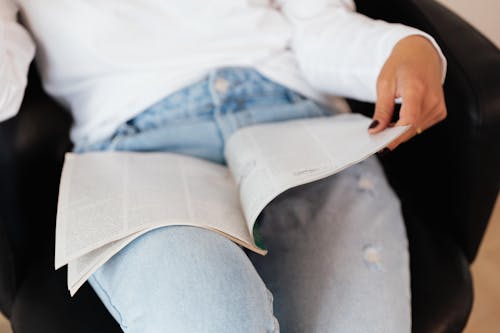 Free Crop unrecognizable lady in jeans and white sweater reading textbook while relaxing in comfortable armchair Stock Photo