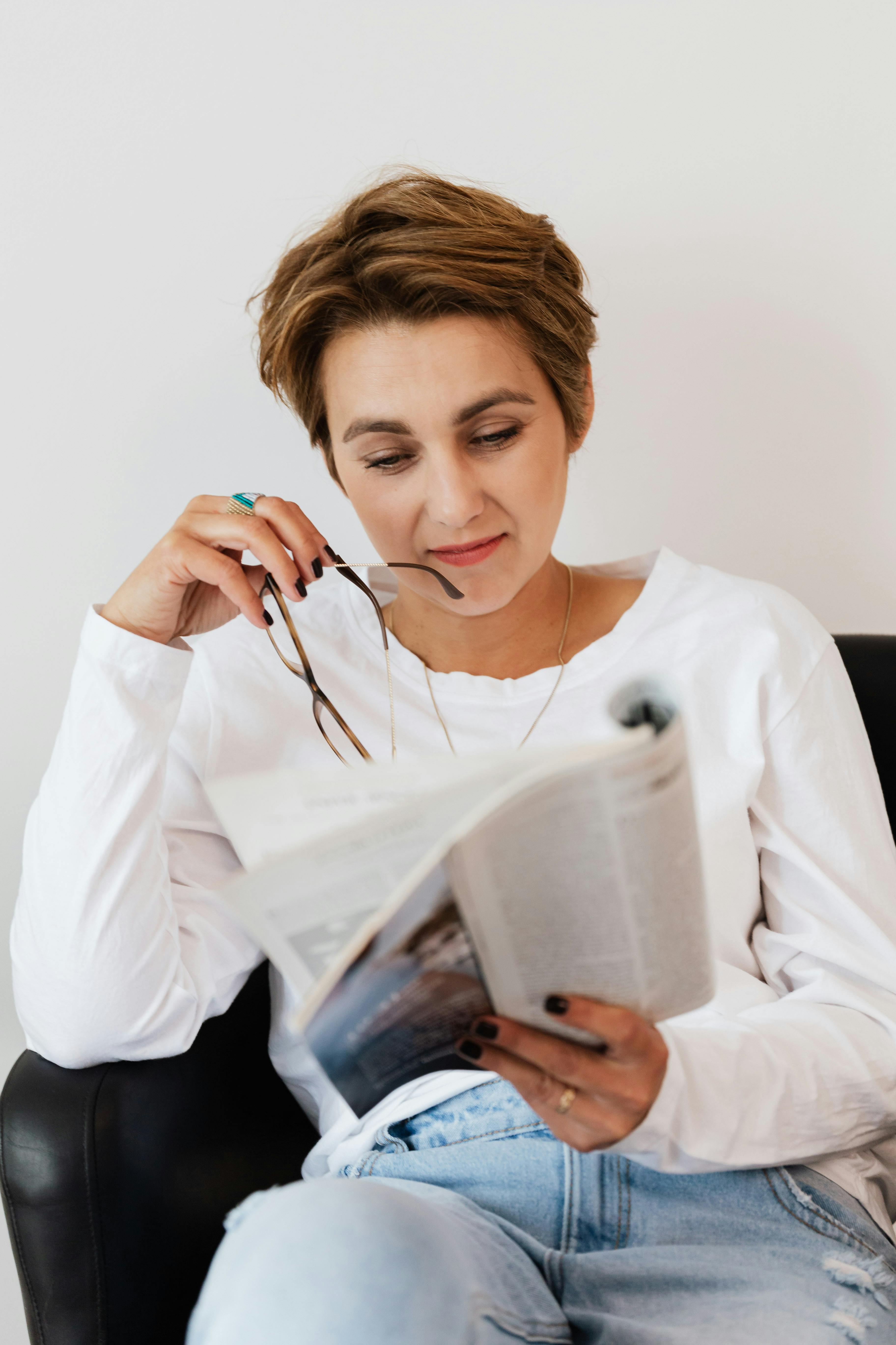 pensive lady with eyeglasses in hand reading magazine in armchair