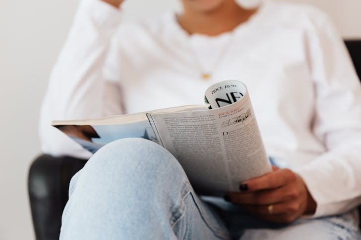Crop unrecognizable female in jeans and white sweater reading magazine sitting in armchair during weekend