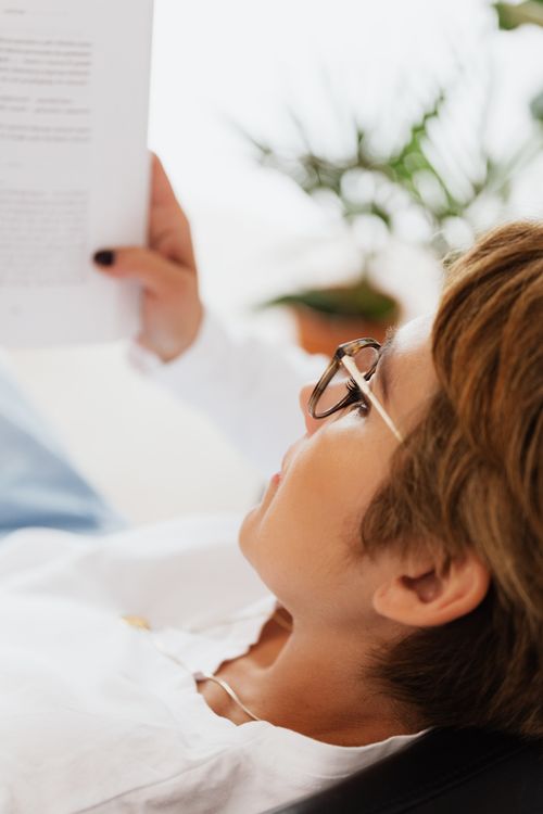 Free Crop female wearing white hoody and eyeglasses lying on comfortable black couch while reading interesting book and relaxing at home Stock Photo