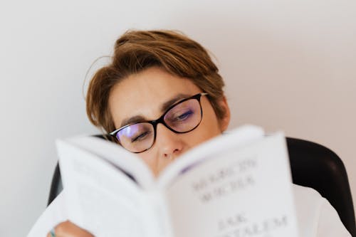 Female wearing glasses reading book while spending free time at weekend at home and searching new information for development