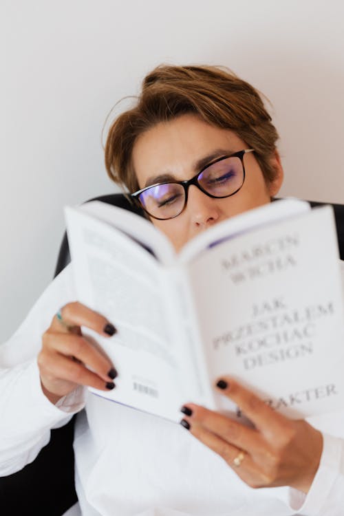 Free Adult female in casual clothes and eyeglasses holding book with white paperback in hands and reading Stock Photo
