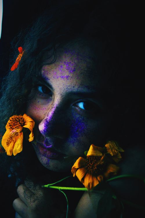Free High angle of young female touching face with delicate orange flowers and looking at camera in darkness Stock Photo
