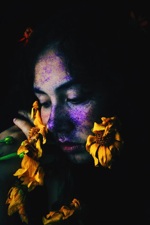 Free Young freckled woman with closed eyes touching face with flowers while resting in darkness Stock Photo