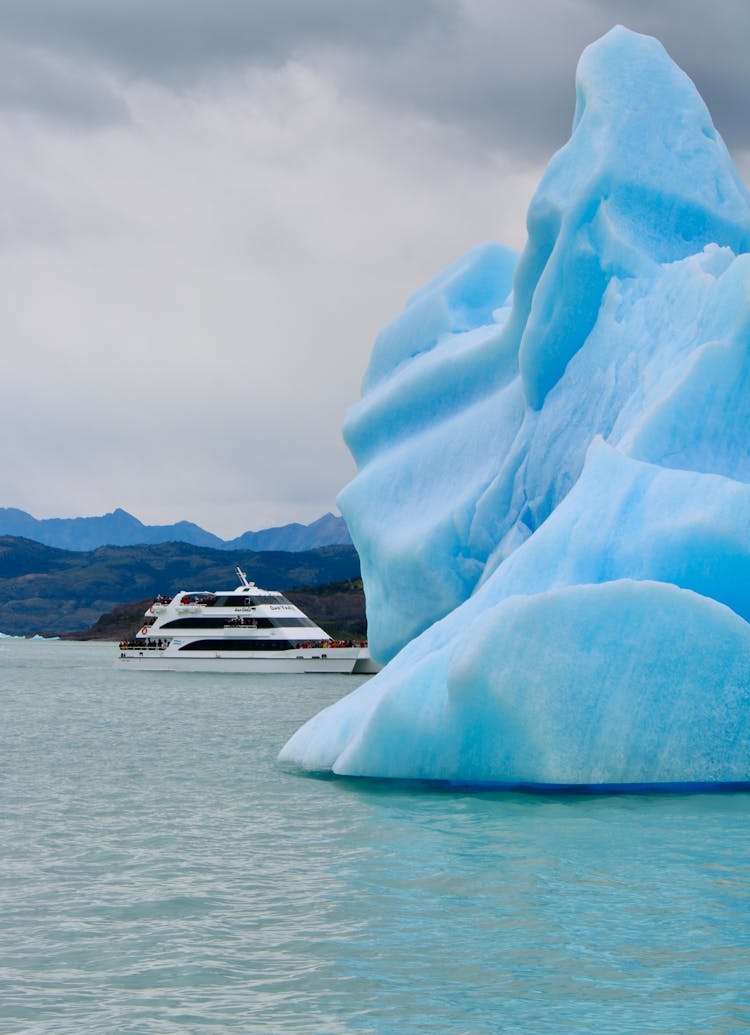 Passenger Vessel Passing By Blue Iceberg Floating On Sea