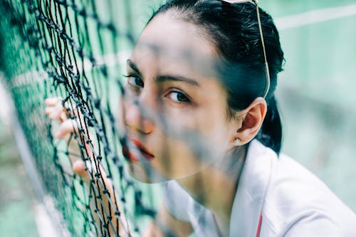 High angle of crop flirting female in t shirt looking at camera through net on tennis court