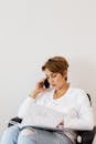 Pensive female in trendy jeans and gold necklace conversing on phone while reading magazine and sitting on chair near white wall