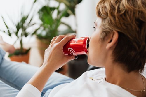 Free Side view of crop anonymous adult dreamy barefoot lady with short hair in jeans lying with crossed legs while drinking coke near potted plants in apartment and looking away Stock Photo