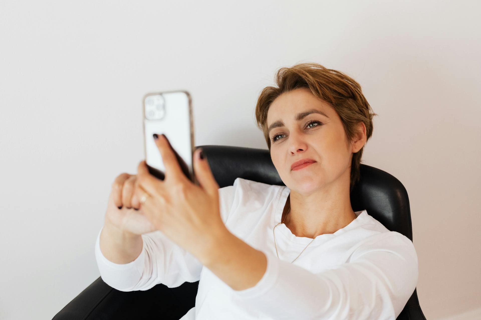 Stylish adult positive female in casual wear with manicure and short hair taking selfie on cellphone while sitting on comfortable leather chair near white wall