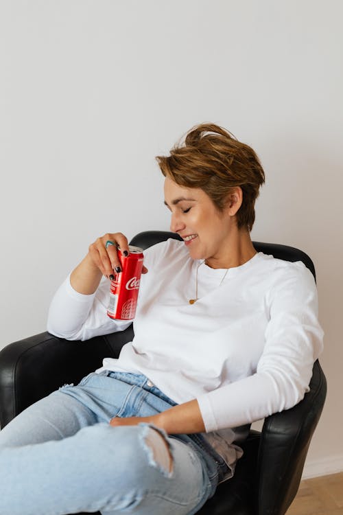 Free Adult smiling woman with short hair in stylish jeans sitting with crossed legs in comfortable leather chair having can of soda Stock Photo