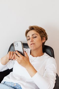 一位穿着牛仔裤和白色衬衫、正在修指甲的体贴女士，坐在白色背景的舒适皮椅上，用手机浏览社交媒体