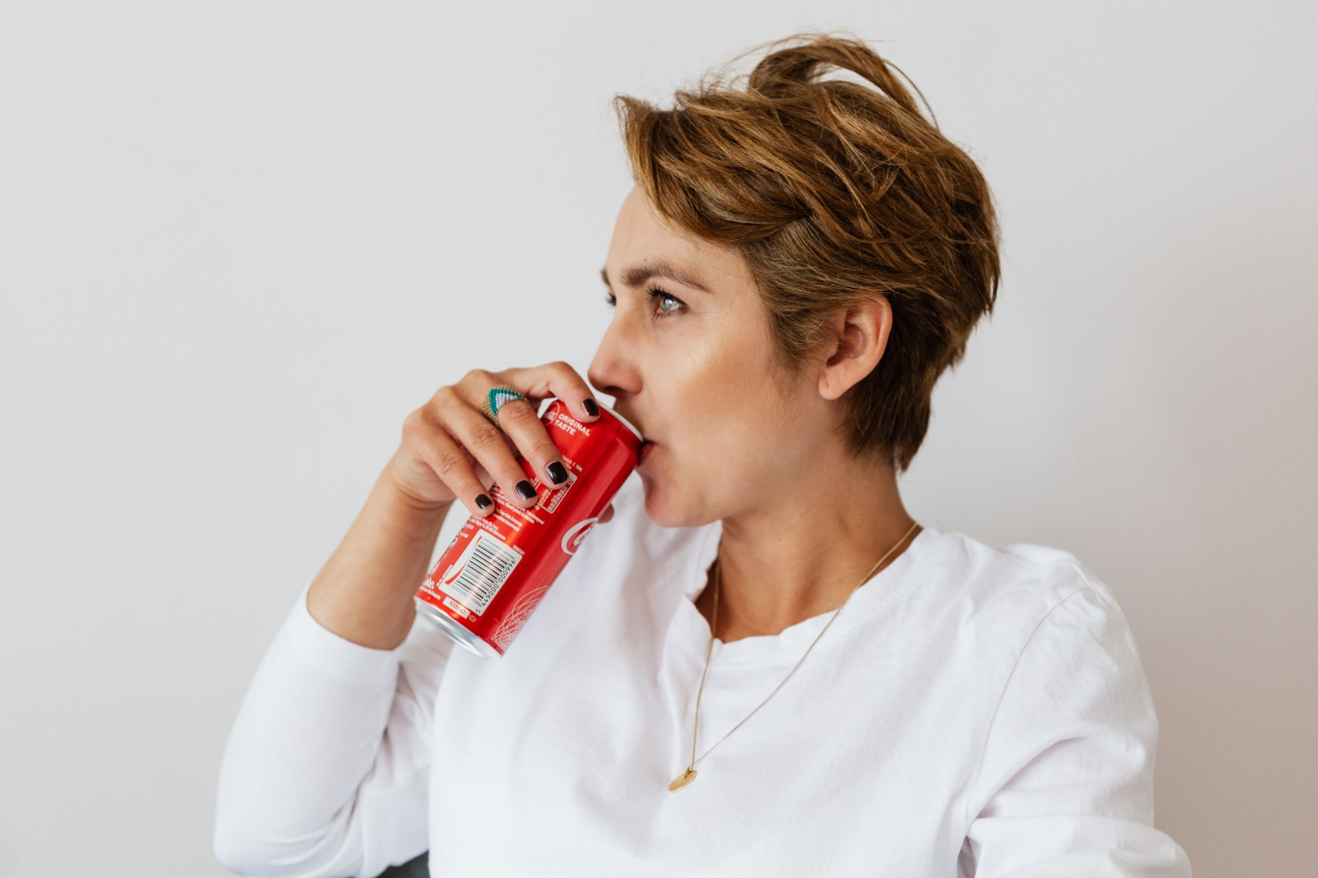 Pensive female in white wear with gold necklace and ring on finger drinking cold coke from red can and looking away